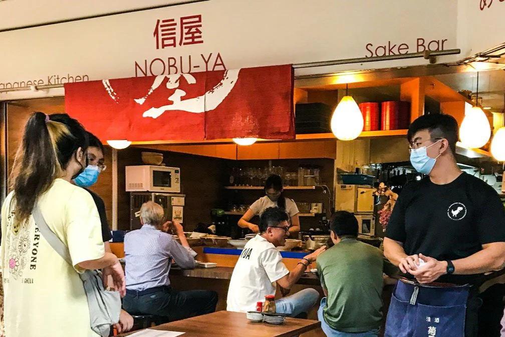 Customers dine and interact at the counter of Nobu-Ya, a Japanese sake bar and kitchen with a warm, casual atmosphere. Staff members wearing masks are visible, including a server in the foreground and a chef preparing food in the background.