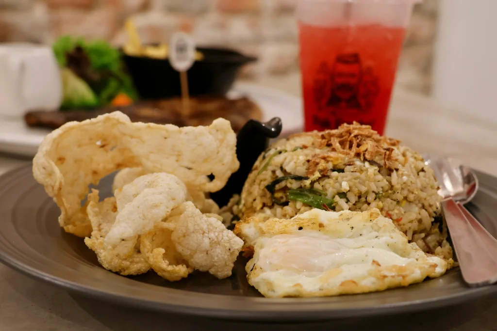 This image displays a plate of Indonesian fried rice, known as Nasi Goreng, topped with fried shallots and served with a fried egg and crispy krupuk crackers. In the background, a steak with fries and a tall glass of bright red iced tea complete the meal setting.