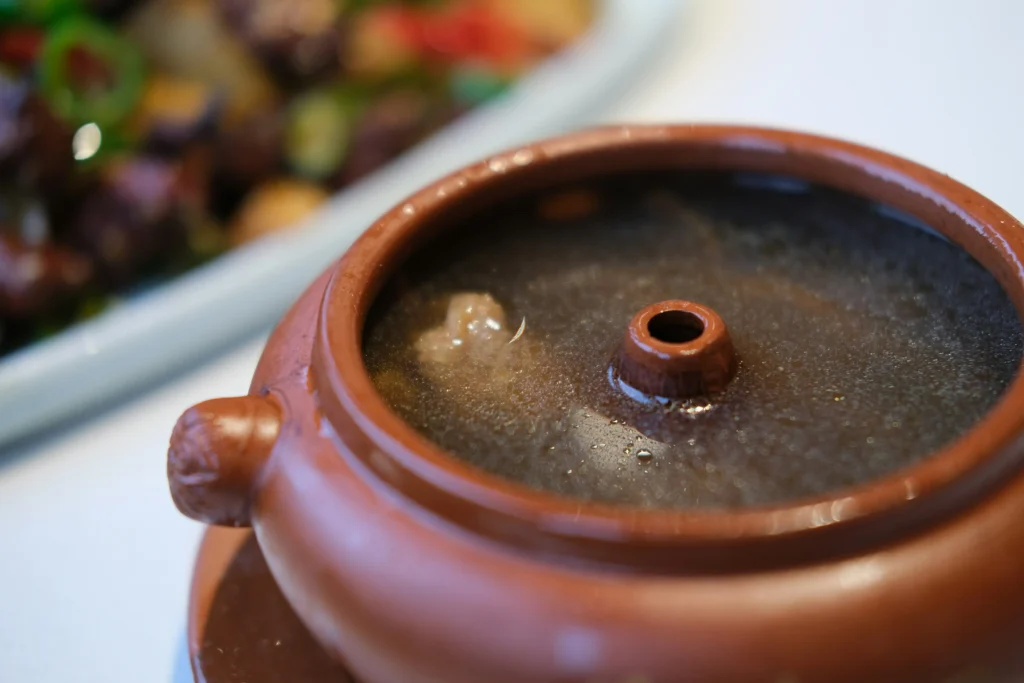 A terracotta steam pot holds a dark, clear broth with visible pieces of meat submerged inside. The unique central chimney of the pot is featured prominently, while a blurred stir-fry dish sits in the background.
