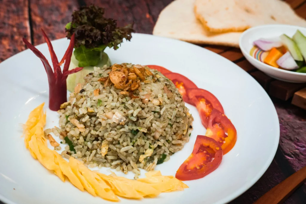 In the image, a mound of seasoned fried rice is elegantly plated with sliced tomatoes, a chili garnish, and shredded egg. The dish is accompanied by side servings of flatbread and a small bowl of pickled vegetables.