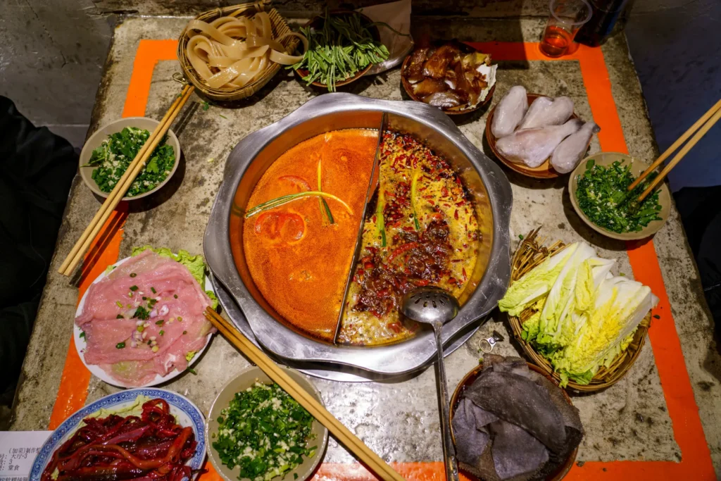 A large metal hot pot sits in the center of a rustic table, featuring a split bowl filled with a creamy orange broth on one side and a dark, spicy chili broth on the other. Surrounding the pot is an array of fresh ingredients, including sliced meats, leafy vegetables, wide noodles, and various bowls of aromatics and dipping sauces.