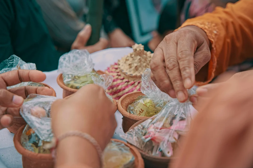 In the image, multiple people reach toward small clay pots filled with plastic-wrapped offerings or treats. A woven, pink-and-tan decorative basket sits in the center of the table amidst the group.
