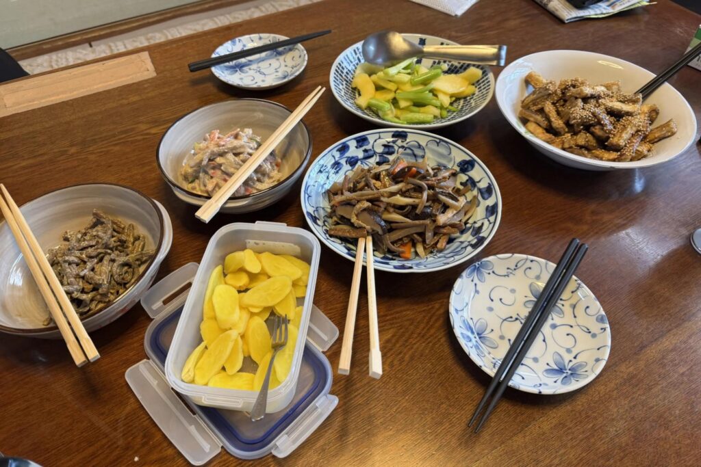 This image displays a wooden dining table spread with several bowls and containers of Japanese-style side dishes, including pickled vegetables and sautéed mushrooms. Multiple sets of chopsticks are placed across the bowls and on patterned small plates, suggesting a communal meal setting.