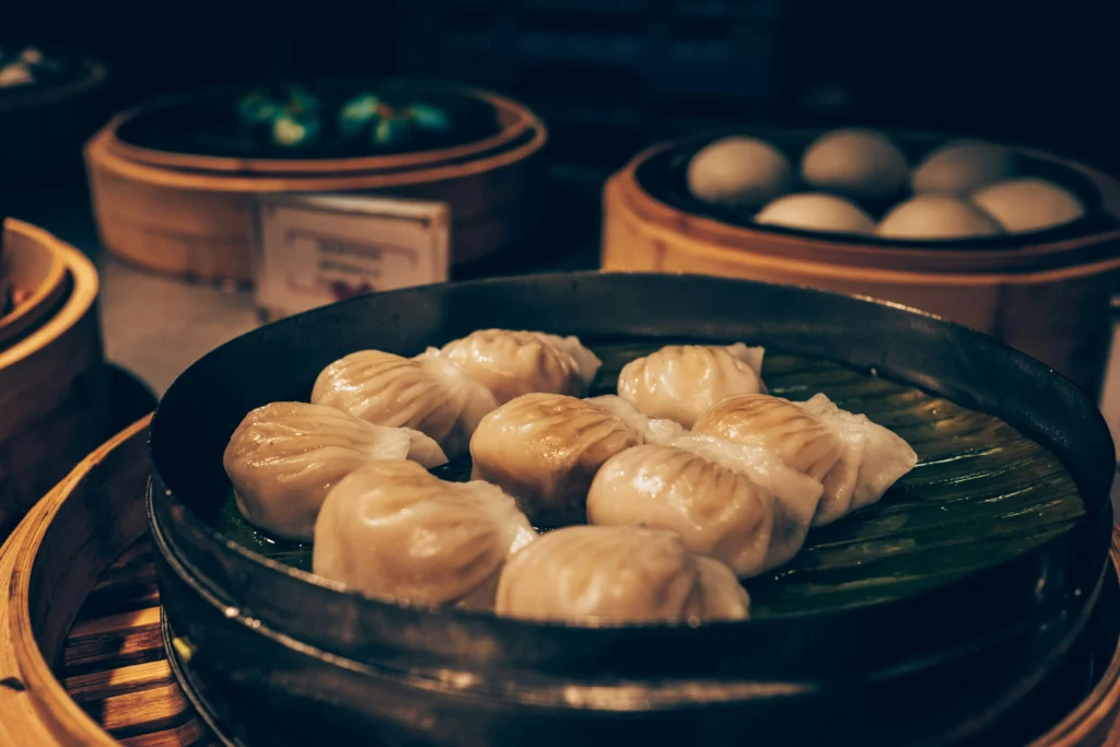 Several bamboo steamers hold a variety of dim sum, including translucent dumplings and soft steamed buns. The foreground focuses on pleated dumplings resting on a green leaf, captured with a moody, cinematic lighting style.