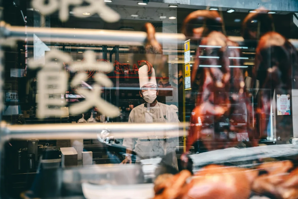 Viewed through a window etched with calligraphy, a masked chef in a tall white hat stands behind a display of roasted ducks. The scene captures the bustling atmosphere of a traditional eatery, framed by a shallow depth of field and warm indoor lighting.