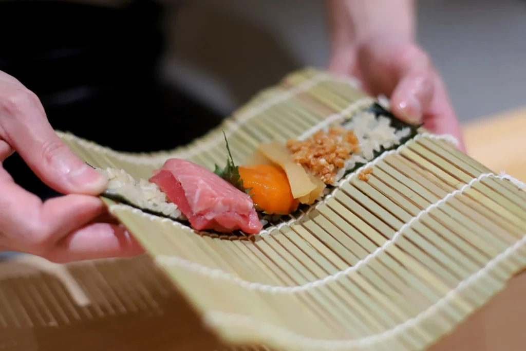 A person's hands hold a bamboo rolling mat containing a piece of nori topped with seasoned rice and an array of colorful sushi ingredients. The fillings include fresh pink tuna, bright orange uni, pickled ginger, a green shiso leaf, and minced toppings lined up for rolling.