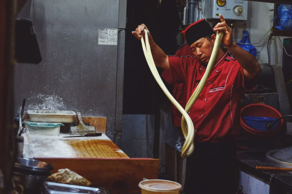 A chef wearing a red uniform and a black hat skillfully pulls and stretches a long loop of dough in a bustling kitchen. The surrounding workspace is dusted with flour and filled with various cooking tools, capturing the traditional art of noodle making.