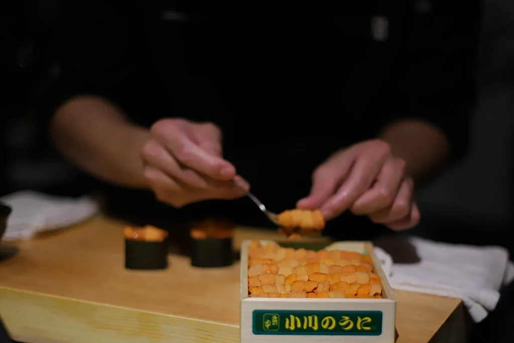 A chef uses a small spoon to carefully lift a piece of bright orange sea urchin (uni) from a wooden box. In the background, two freshly prepared gunkan maki sushi rolls sit on a light wooden counter.