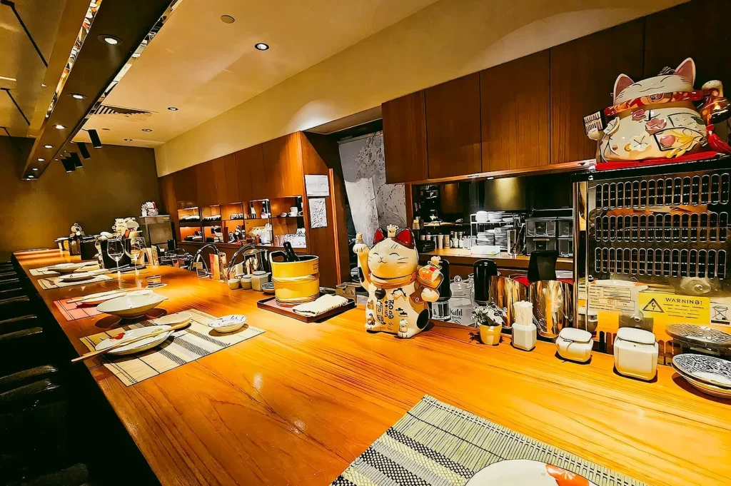 Elegant sushi counter at Hanashizuku Japanese Cuisine Singapore with polished wooden bar, open kitchen, and welcoming lucky cat décor.