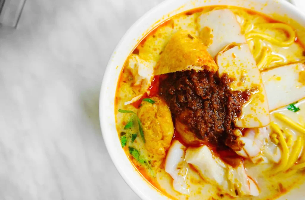 Bowl of spicy curry laksa with noodles, fish cake, and sambal, highlighting iconic Malaysian-Singaporean comfort food.