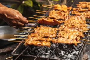 Thai street vendor grilling moo ping pork skewers over charcoal with bamboo sticks and wood smoke