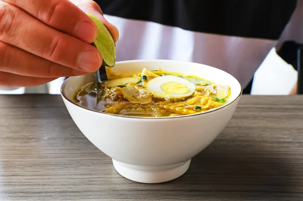 Hand squeezing fresh calamansi over a steaming bowl of noodle soup, adding citrus flavor to a classic Singapore hawker noodle dish.