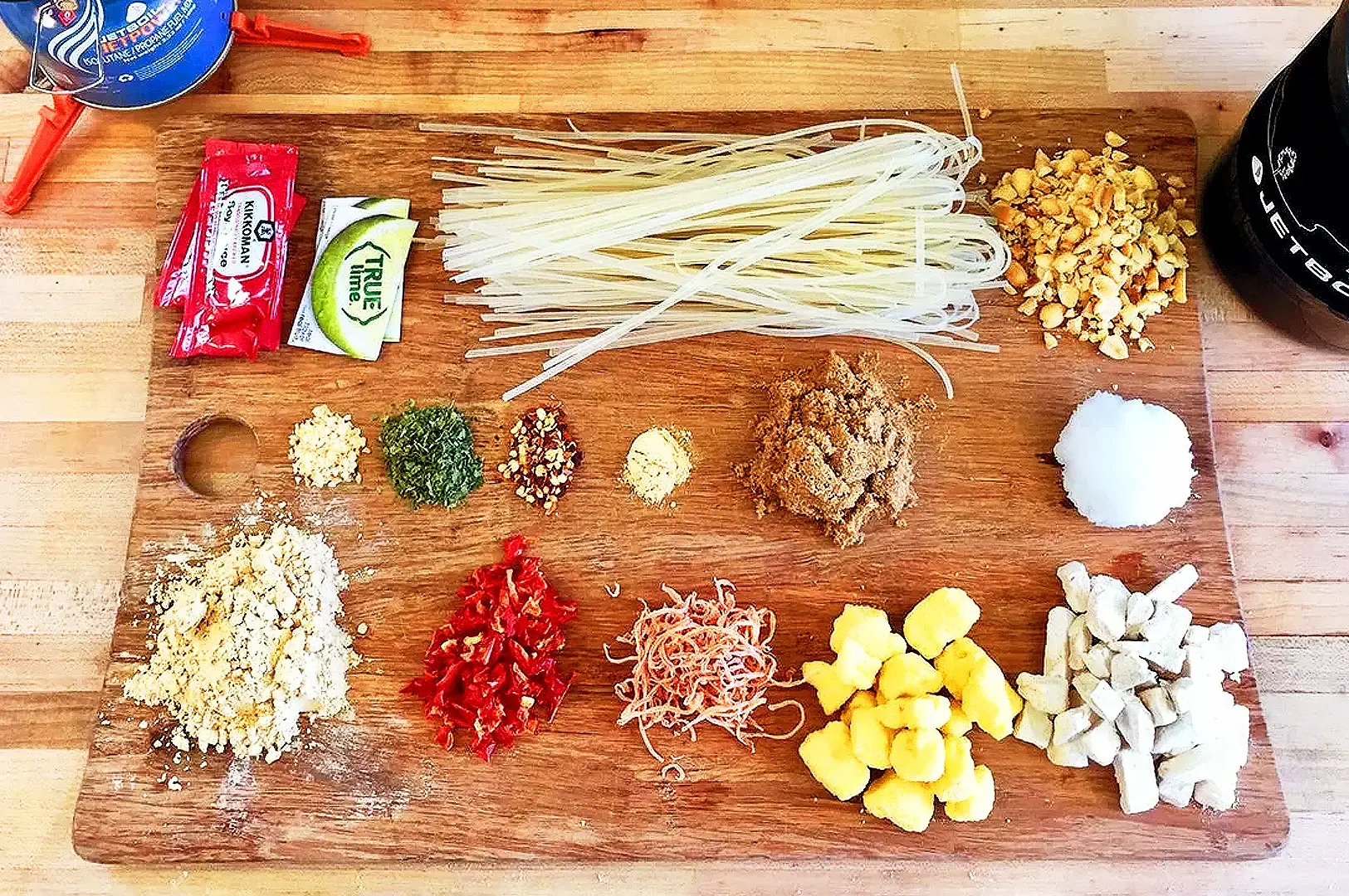 Pad Thai recipe ingredients arranged on a wooden board, including rice noodles, peanuts, chili, dried shrimp, lime and Thai seasonings