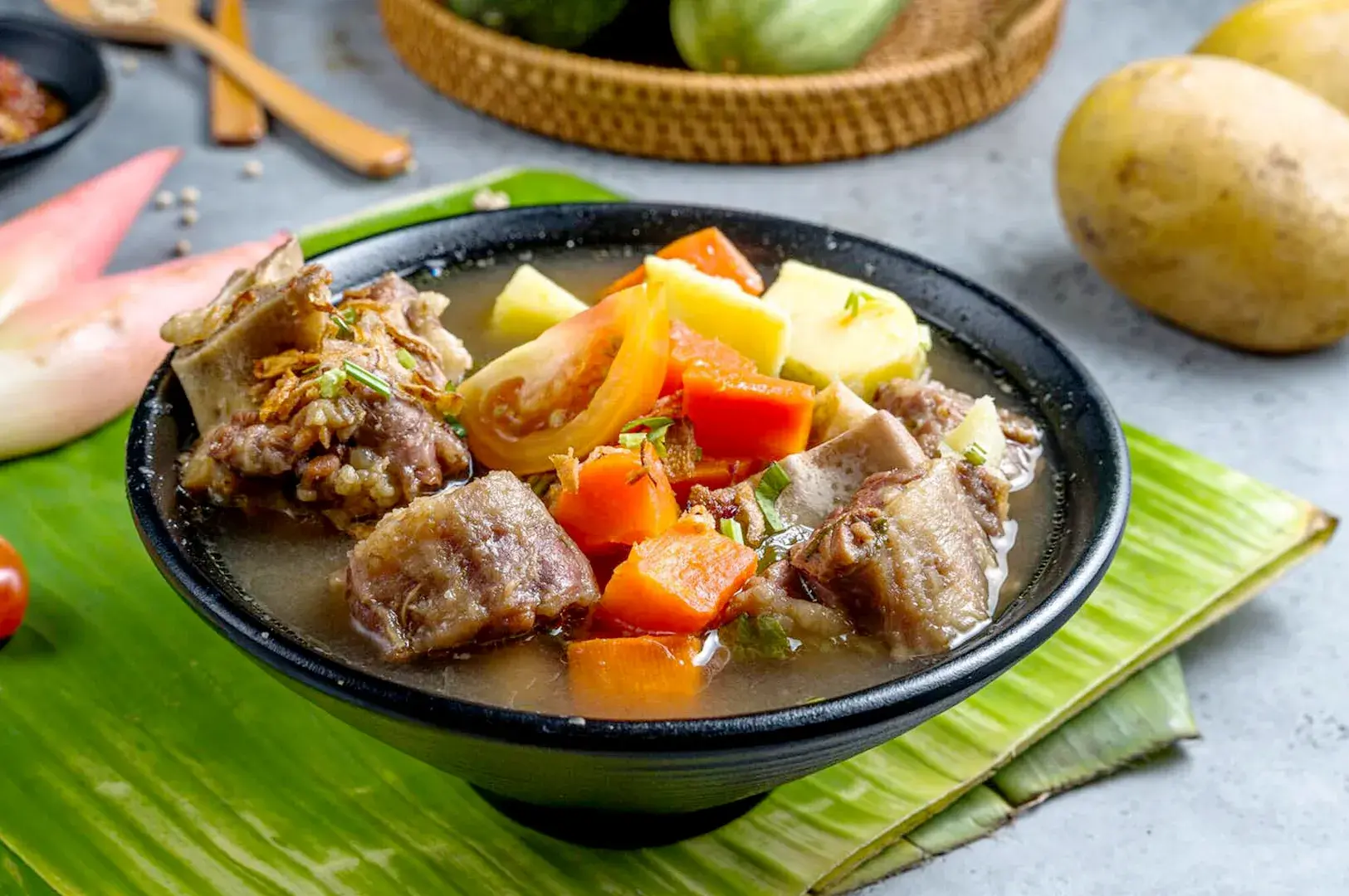 Indonesian-style fried duck with vegetables, potatoes, carrots, tomatoes, and savory broth served in a black bowl on a banana leaf.