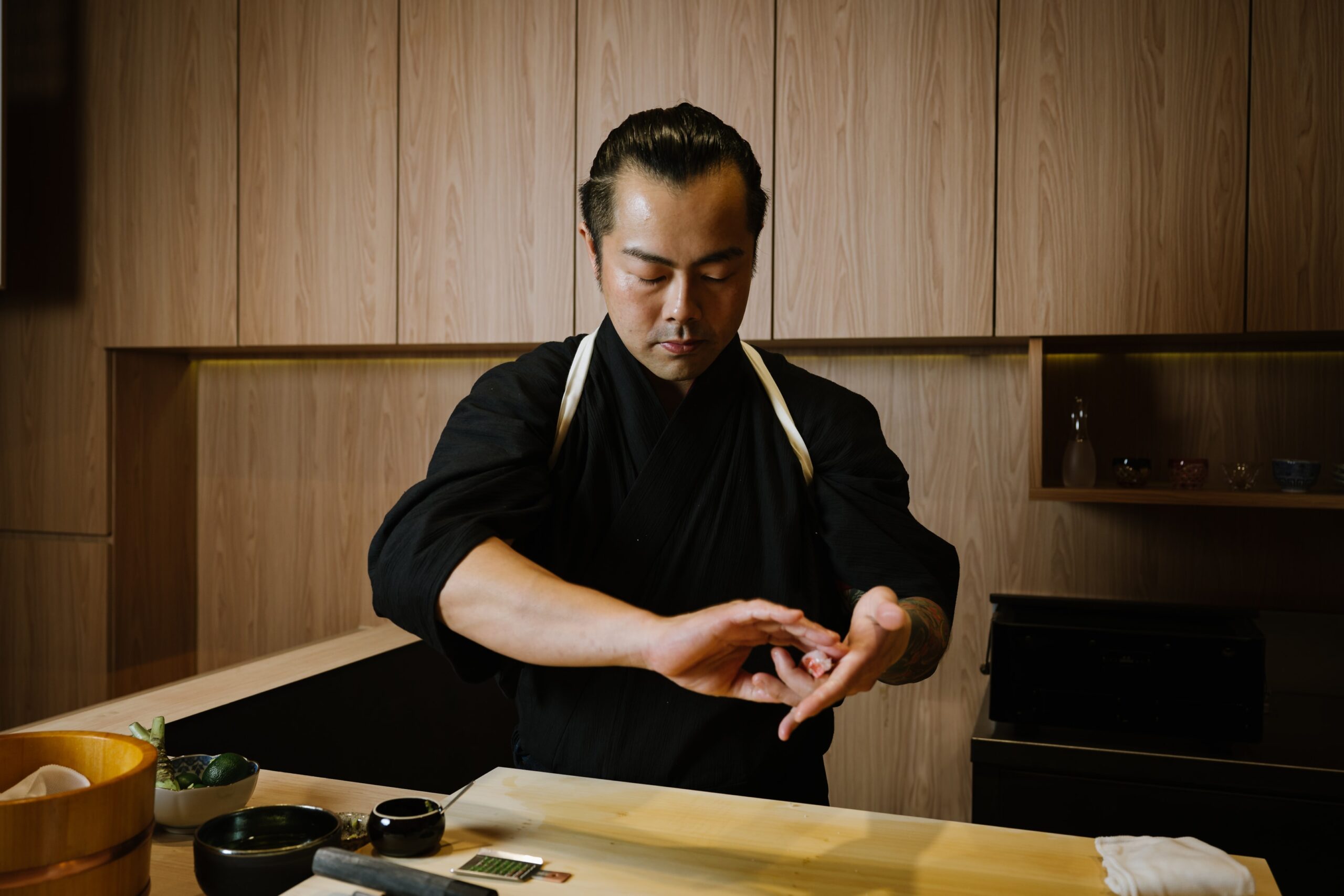 Chef Masa in a black uniform shaping sushi by hand at a wooden counter, with utensils and ingredients arranged in front of him.
