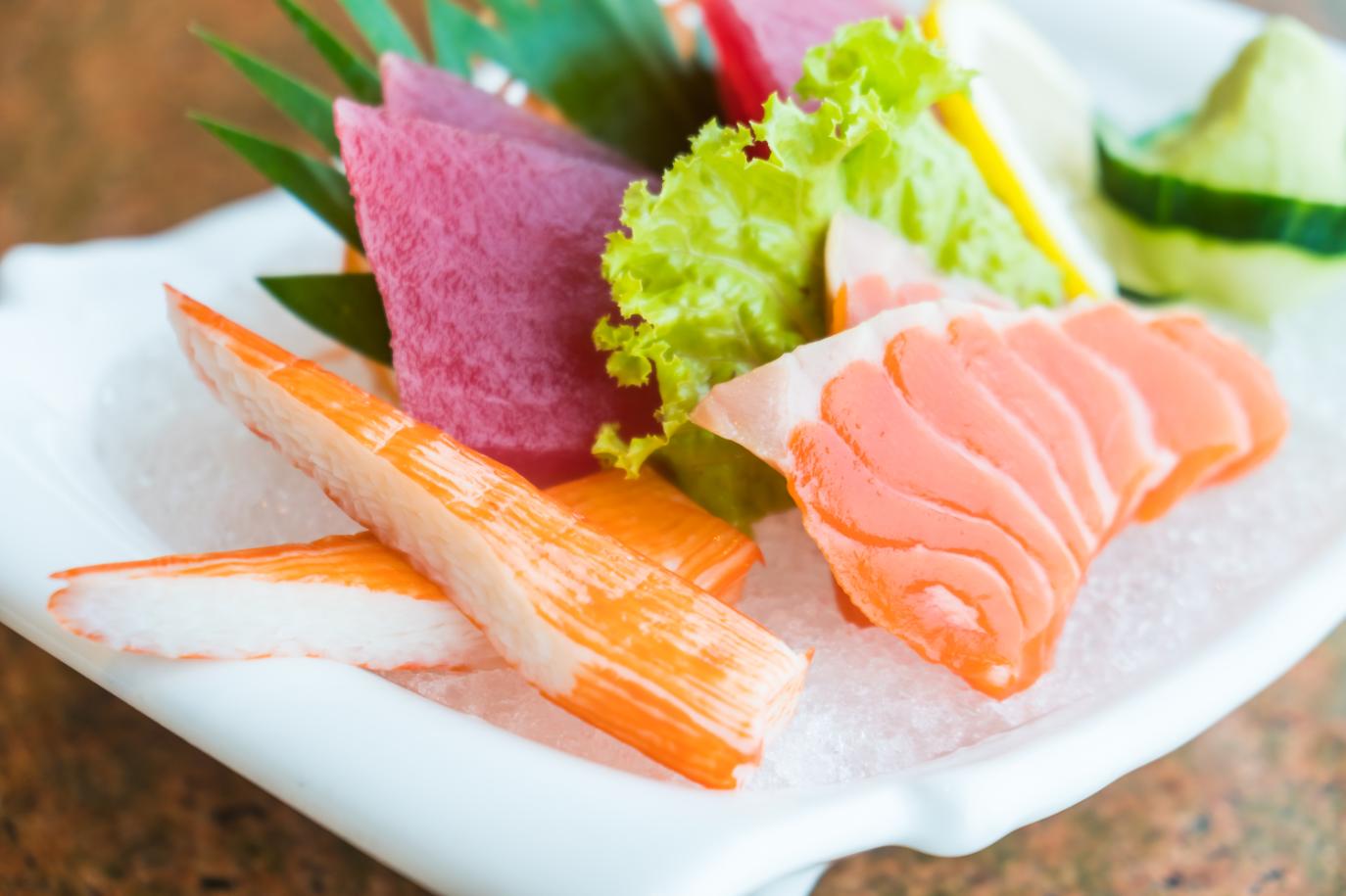 Plate of assorted sashimi on ice, featuring slices of salmon, tuna, imitation crab sticks, and leafy garnish.