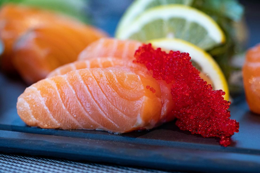 Close-up of fresh salmon sashimi topped with bright red fish roe, served with lemon slices and greens in the background.