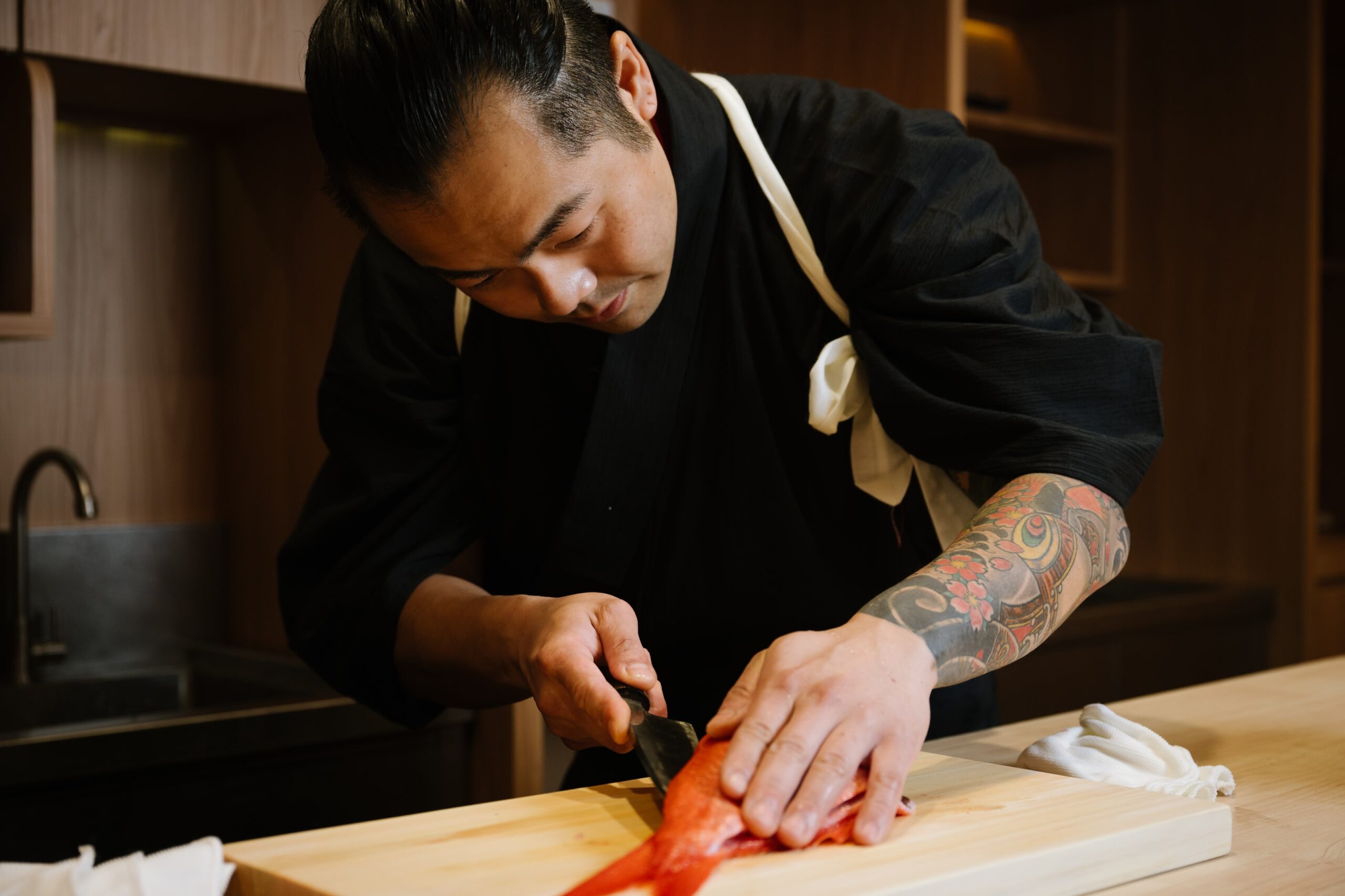 Chef Masa in a black uniform carefully filleting a red fish on a wooden cutting board, showing focused technique and tattooed forearm.