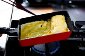 Tamagoyaki being rolled in a traditional Japanese rectangular pan over a gas stove